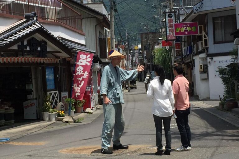 A man in traditional Japanese clothing and hat is giving directions to two tourists on a street in Japan.