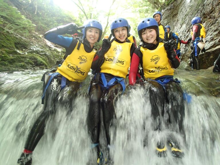 Three women in wetsuits and helmets sit on a waterfall, smiling and giving peace signs, while others stand behind them.
