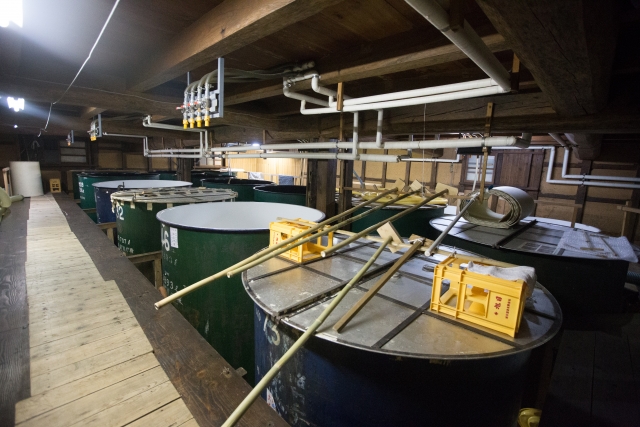 A low-lit fermentation room with large vats, wooden paddles, and wooden crates on top.
