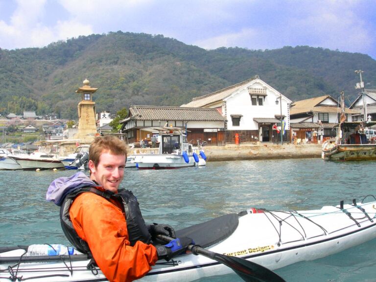A smiling man in a kayak is paddling on the water with Japanese buildings and mountains in the background.