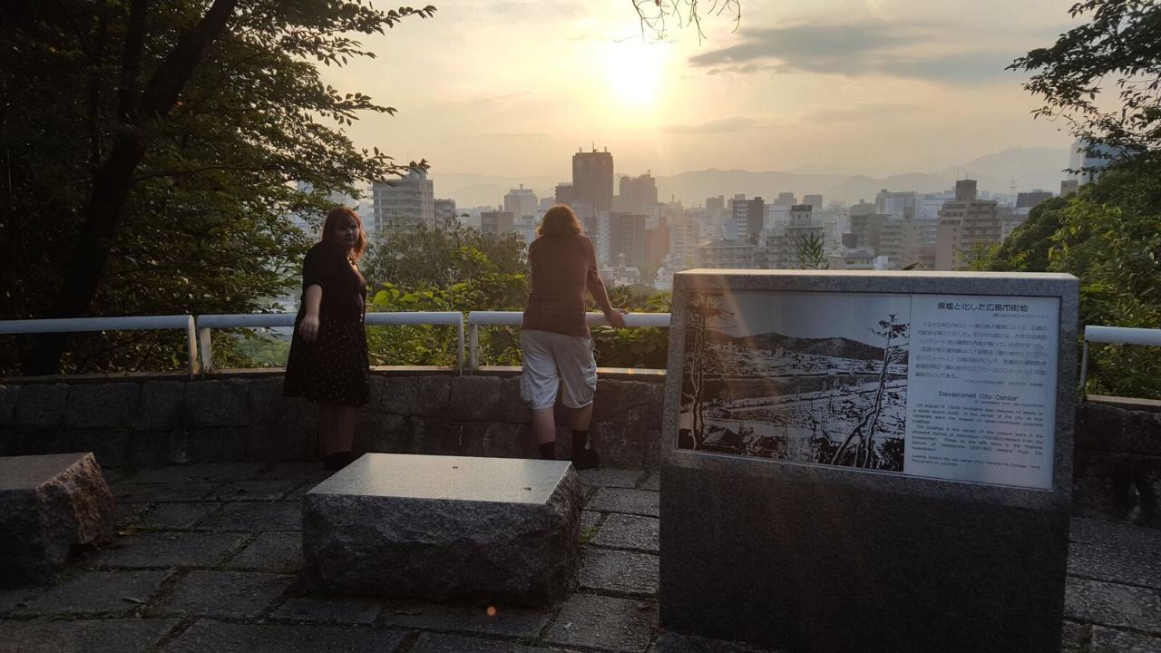 Two people stand at a viewing platform, observing the city skyline at sunset, with a memorial stone featuring text and images beside them.