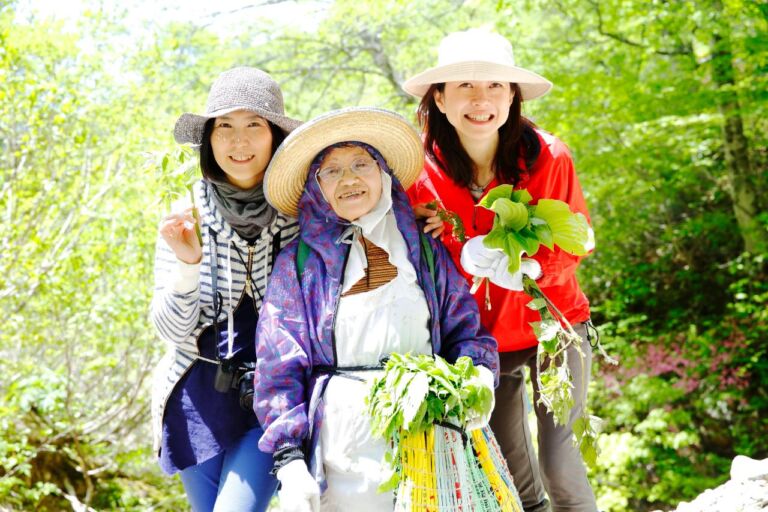 Three Asian women are posing outdoors surrounded by green foliage and trees. They are all wearing hats and holding greenery or a basket filled with green plants.