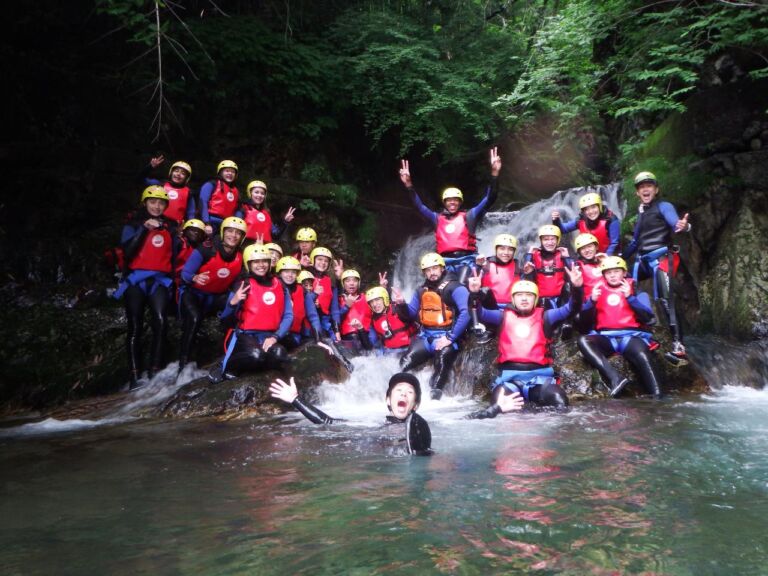 A large group of people in helmets and life vests pose for a photo in a river near a waterfall, some sitting on rocks and one person in the water.