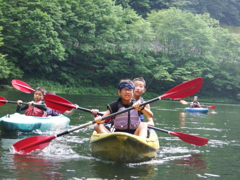 Several children are kayaking on a lake surrounded by lush green trees.