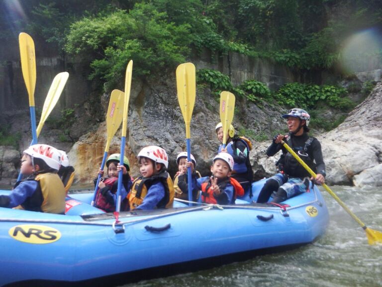 A group of children and an adult are enjoying a whitewater rafting trip on a blue NRS raft. They are wearing helmets and life jackets, and are equipped with paddles.