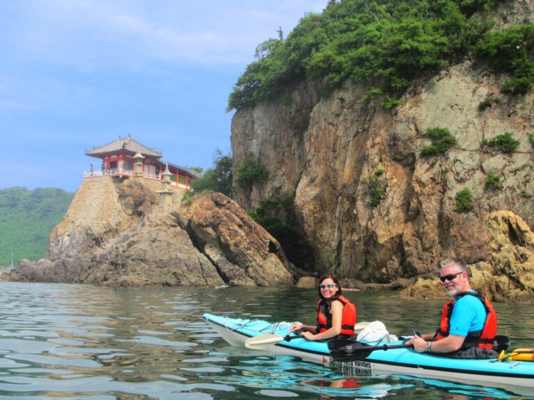 A couple wearing life vests is kayaking near a rocky cliff with a traditional Japanese building on top.