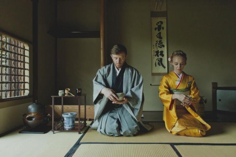 A man and a woman dressed in traditional Japanese clothing kneel in a tatami room, participating in a tea ceremony.