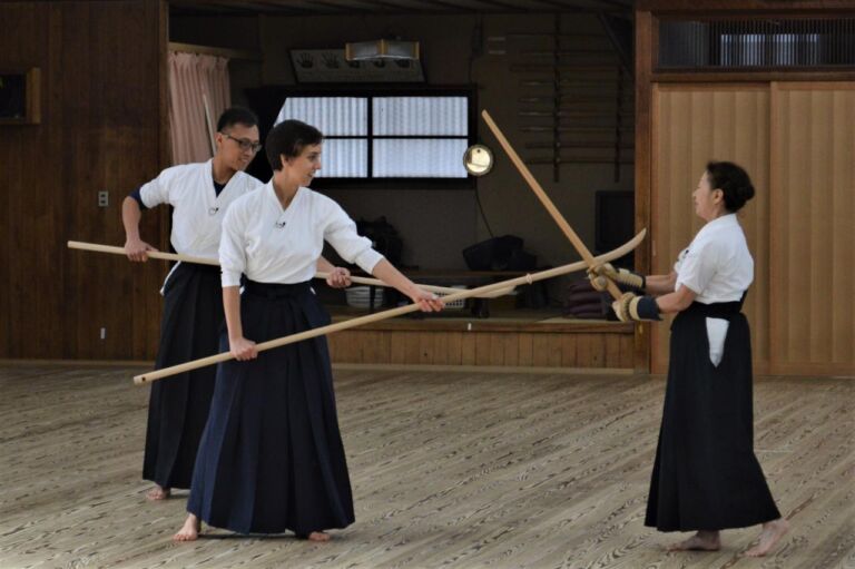 Three people in traditional Japanese clothing are engaged in a martial art practice, with two holding wooden poles and the third wielding a wooden sword, on a wooden floor in a dojo-like setting.