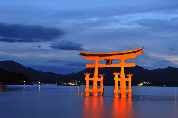 An orange torii gate stands in the water at dusk with mountains and a cloudy sky in the background.