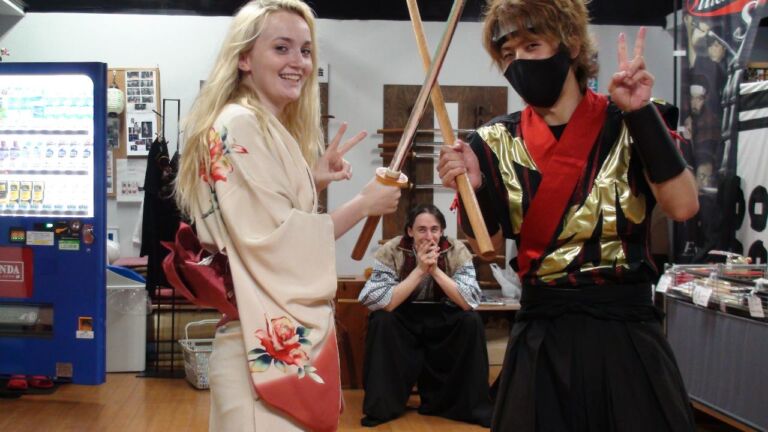 Three people pose with wooden swords in what appears to be a souvenir shop. A woman in a kimono and a man in a ninja costume are making peace signs while another man sits behind them with his hands clasped.