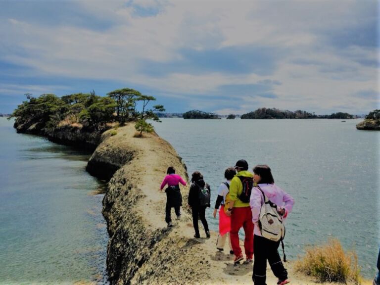 A group of people walks along a narrow strip of land connecting to a small island with trees, surrounded by water and under a cloudy sky.